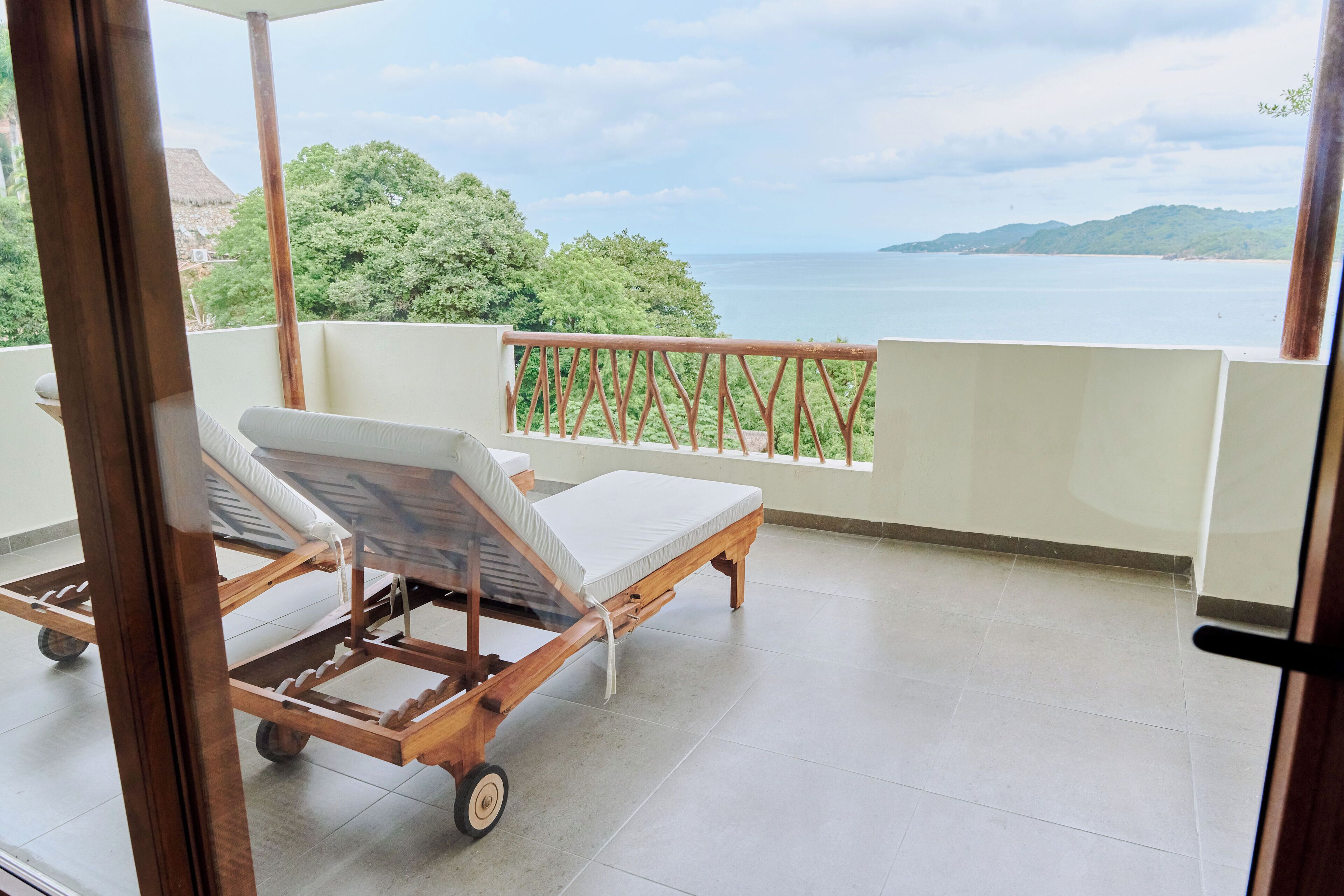 A balcony with a wooden lounge chair and a small table. The balcony overlooks a body of water and greenery.