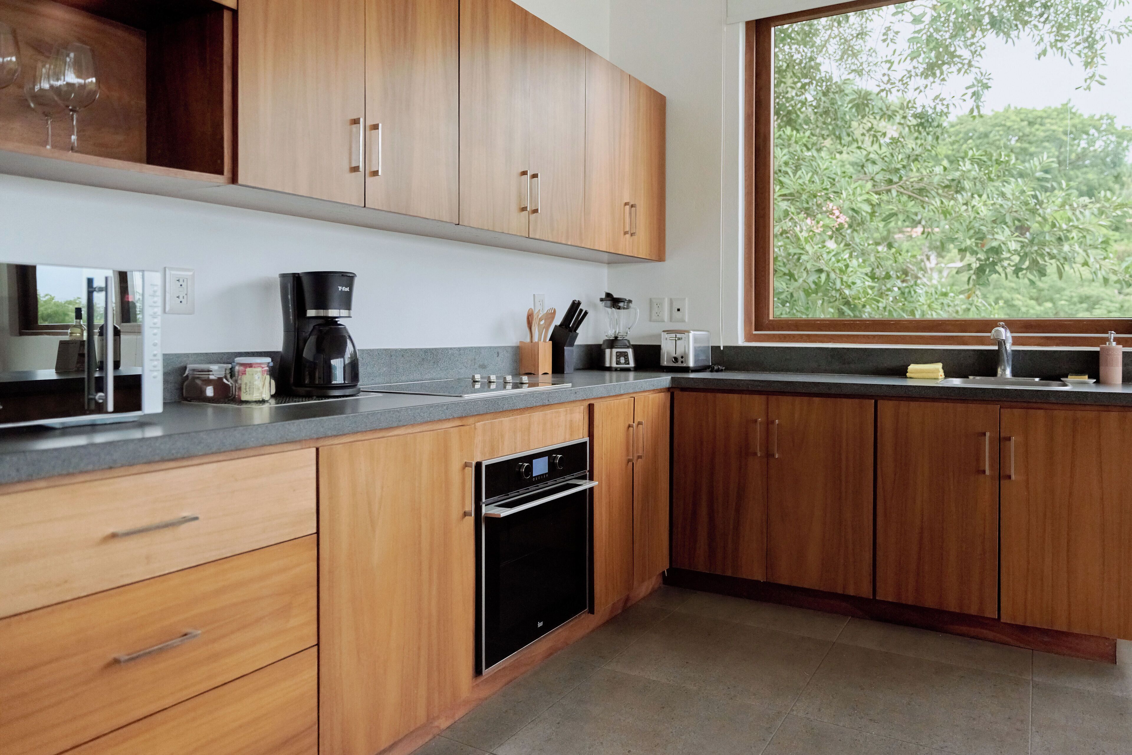Een moderne keuken met houten kastjes, een inbouwoven, een koffiezetapparaat en een raam met uitzicht op bomen.