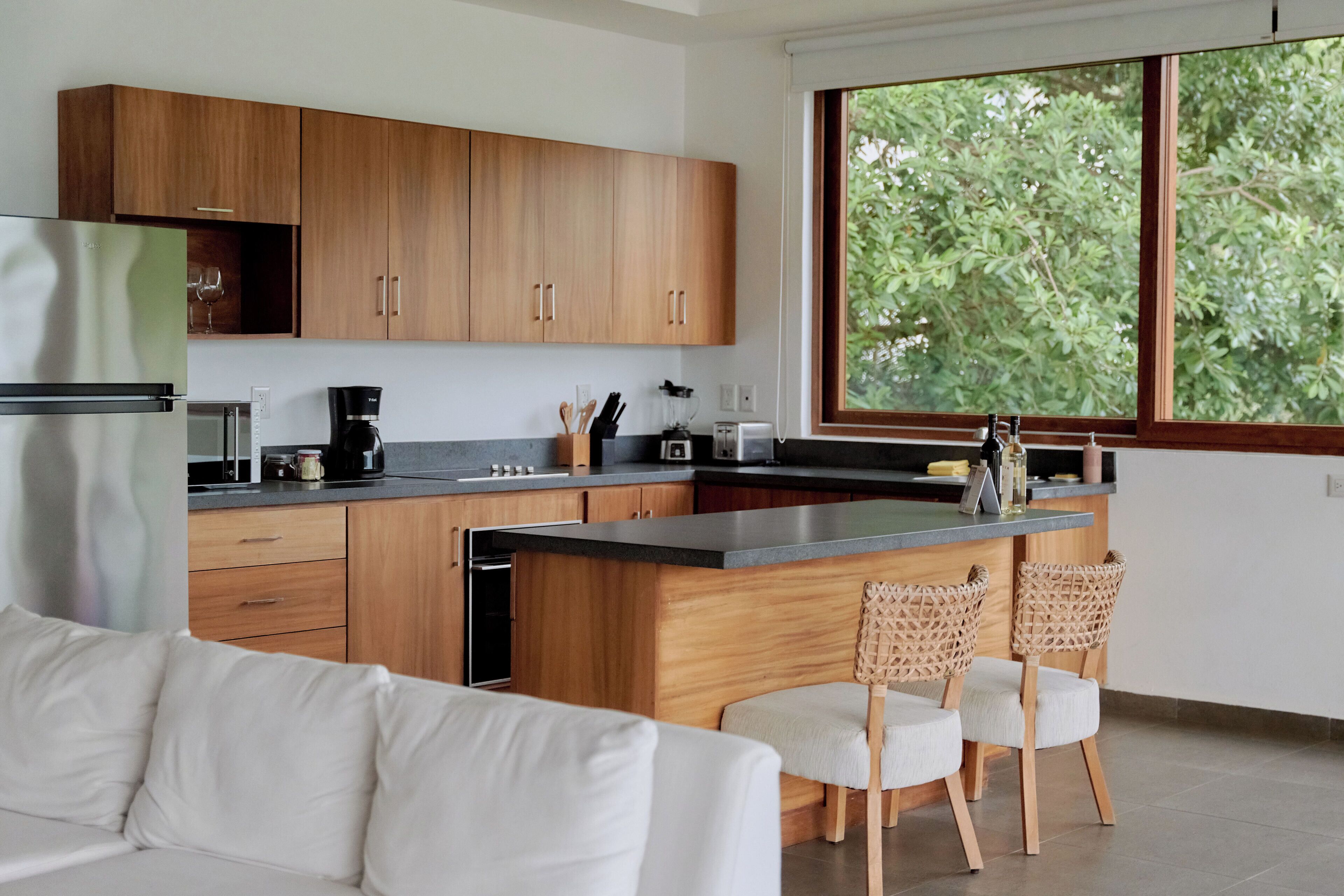 A modern kitchen with wooden cabinets, a large window, and a bar area with two wicker chairs.