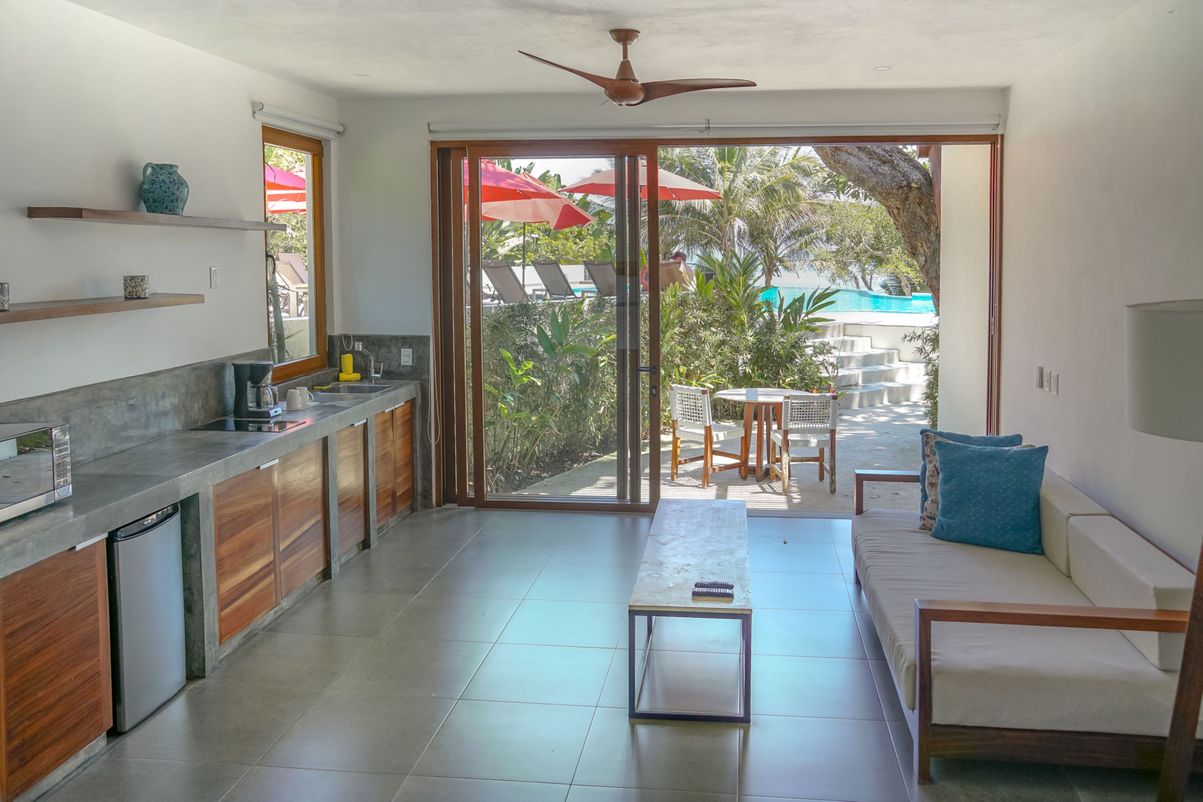 A modern kitchen with a built-in dishwasher, wooden cabinets, and a large sliding glass door leading to an outdoor seating area with a pool and umbrellas.