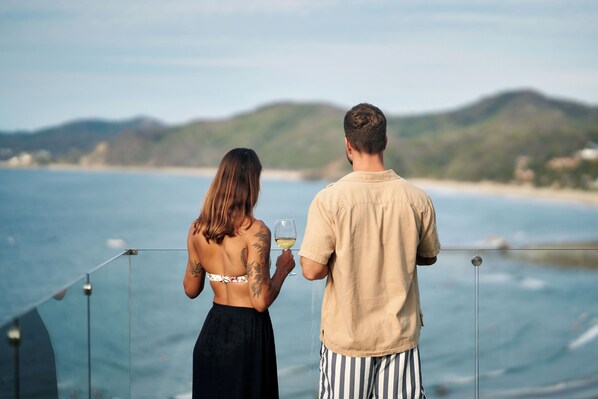 A couple standing by a railing overlooking a body of water with hills in the background.