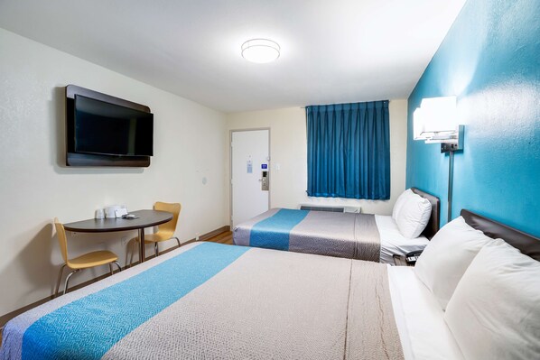Habitación de hotel con dos camas, un escritorio con sillas, un televisor de pantalla plana montado en la pared y una ventana con cortinas.