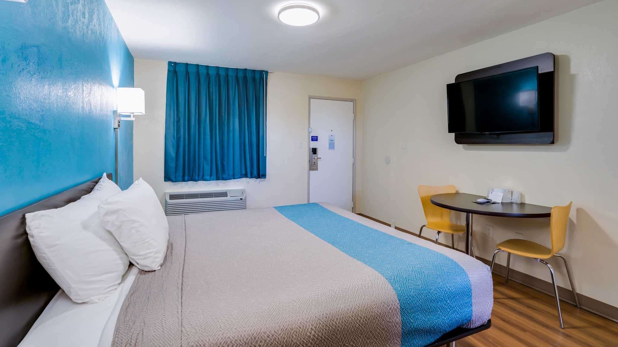 A hotel room with a bed, a desk, two chairs, a television, and a window with blue curtains.