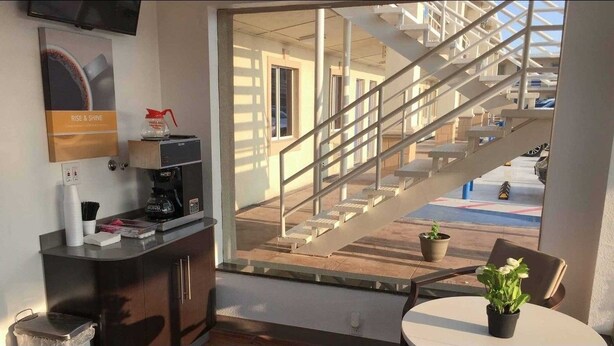 A hotel room with a coffee machine, a trash bin, a small table with a potted plant, and a view of a staircase leading to an outdoor area. - Motel 6 Odessa, TX - 2nd Street (Odessa)