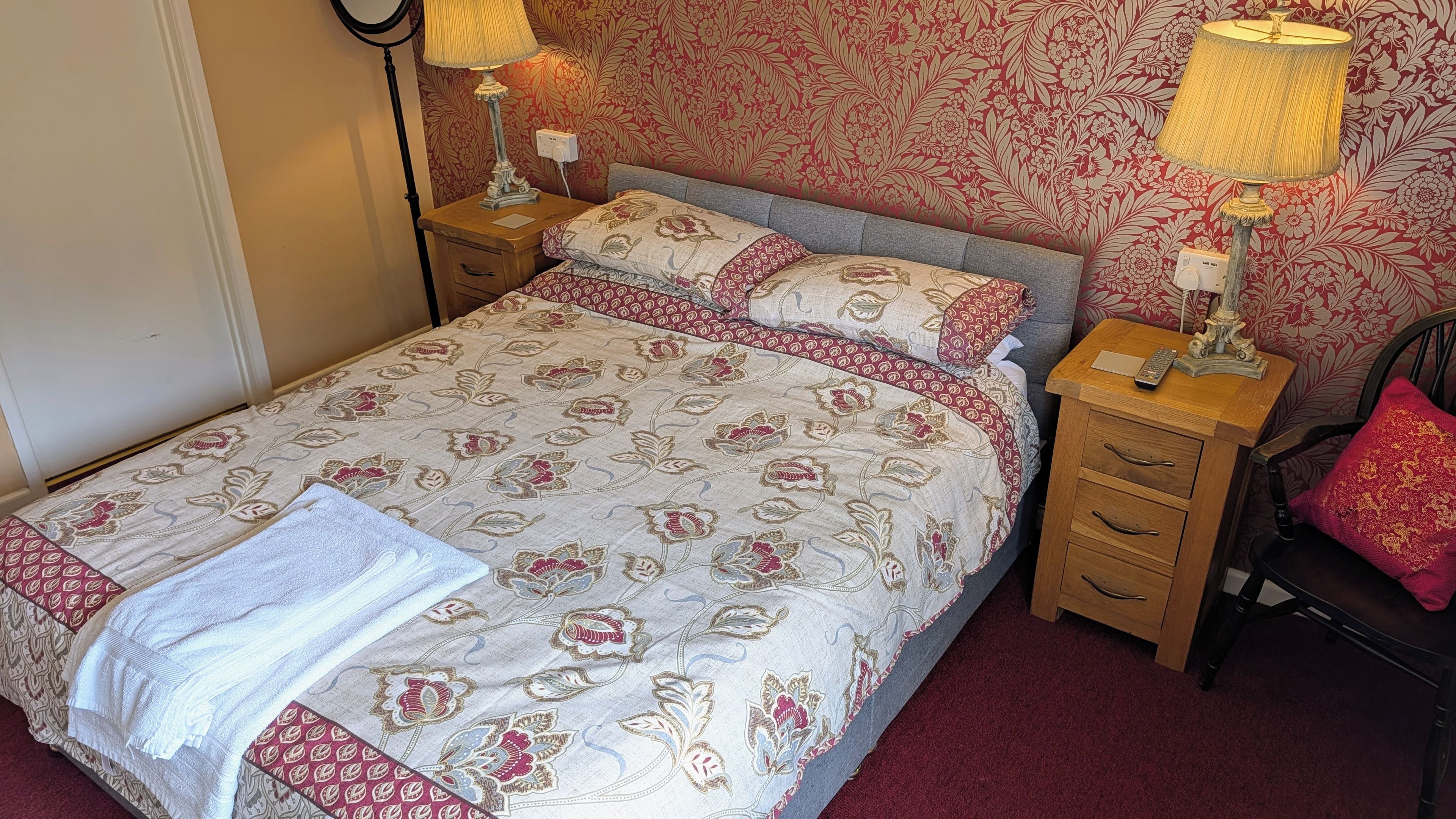 Una cama bien hecha con ropa de cama floral, dos lámparas de noche, una mesita de noche de madera y una silla roja.