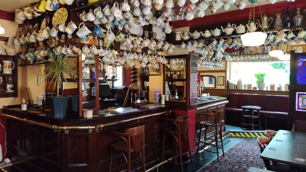 A bar area with a variety of teapots and cups hanging from the ceiling, wooden bar counter, stools, and a large mirror. - The Bull (Birmingham)