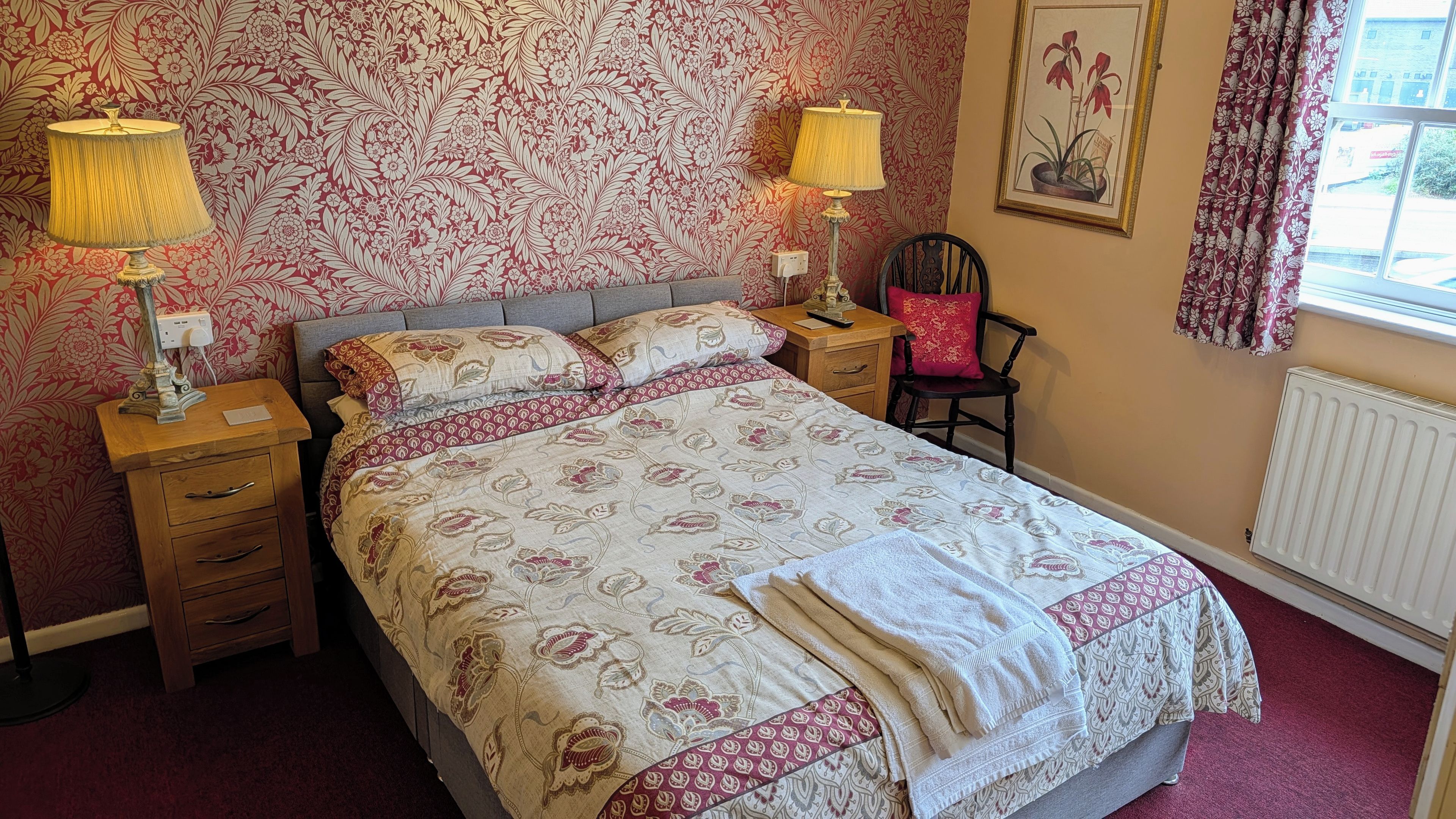 A bedroom with a bed, two bedside tables with lamps, a chair, a radiator, and a window with curtains.