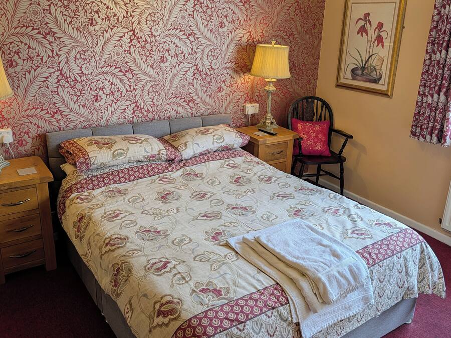 Un dormitorio con cama, dos mesitas de noche con lámparas, una silla, un radiador y una ventana con cortinas.