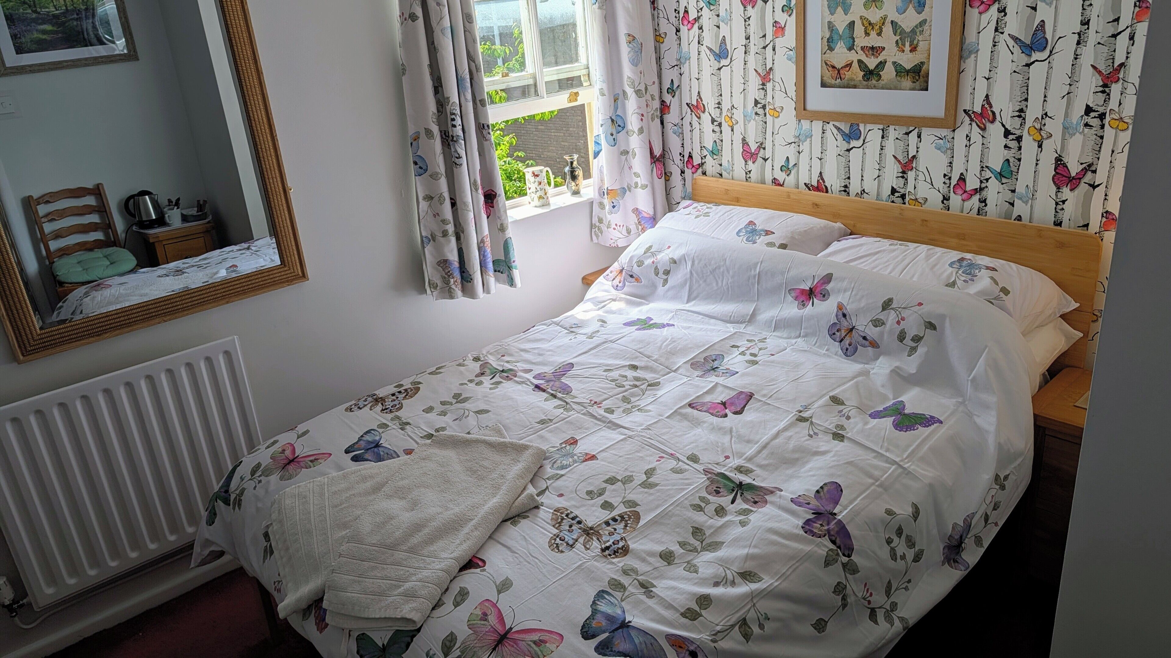 Un dormitorio con cama, un radiador, una ventana con cortinas, un espejo y un papel tapiz de mariposas en la pared.