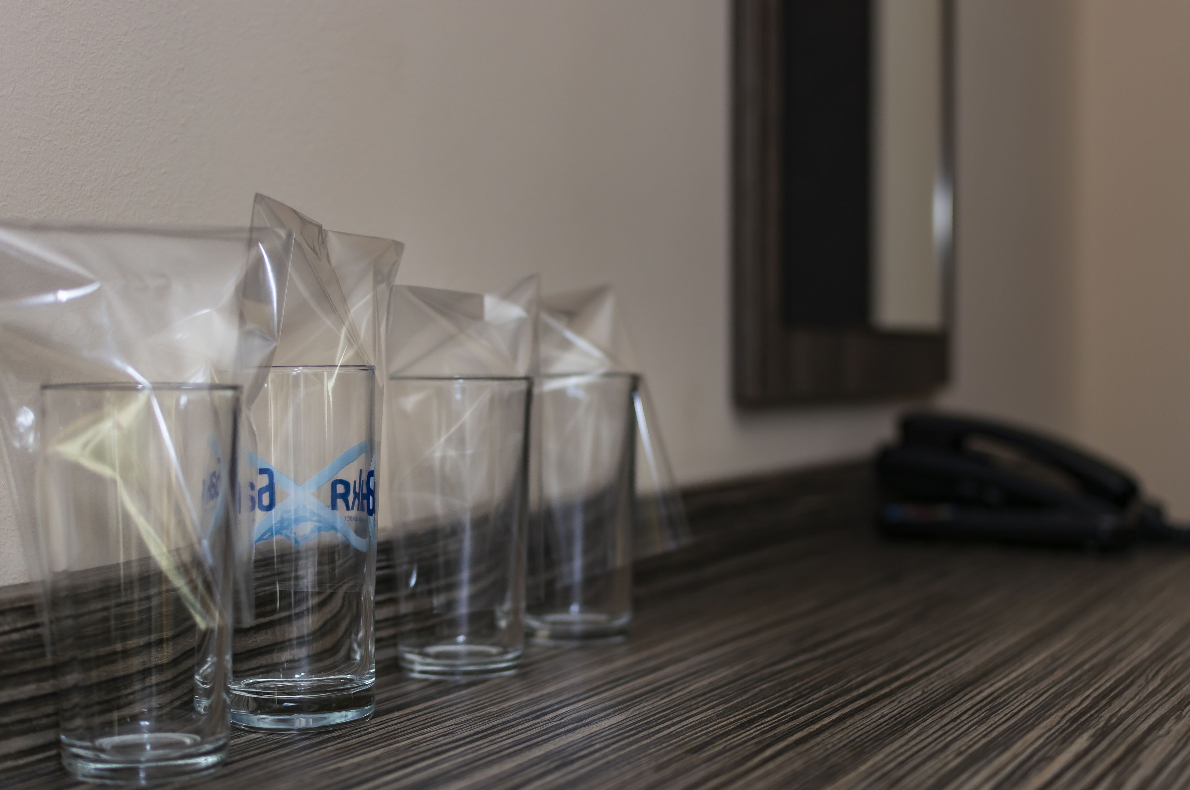 A hotel room with a wooden table, four clear glasses with a logo, and a black telephone.