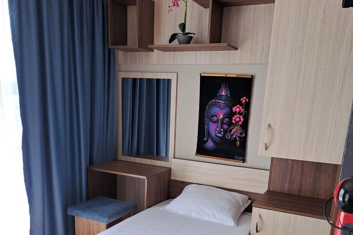 A hotel room with a bed, a desk, a chair, a shelf with a potted plant, and a framed artwork on the wall.