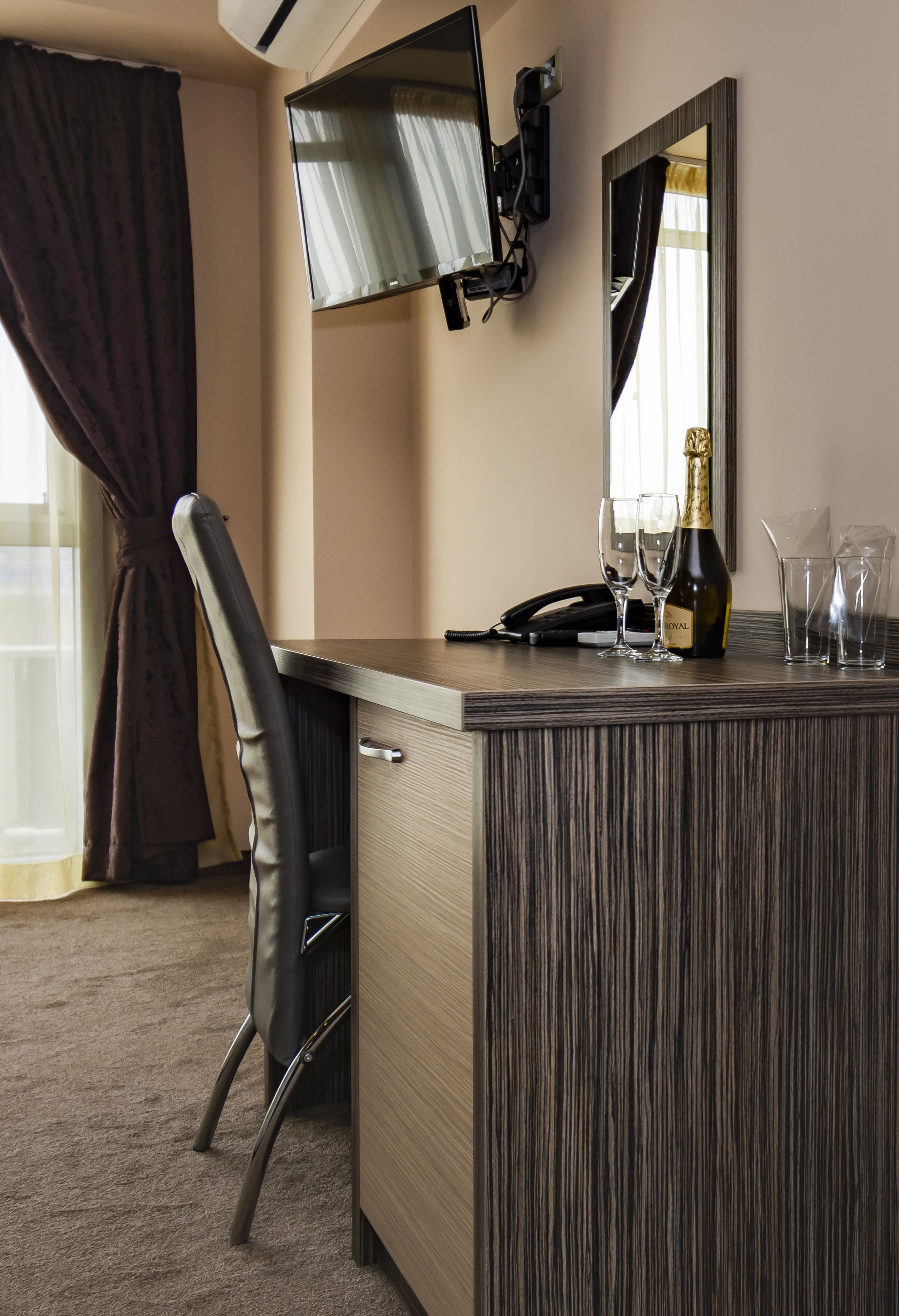 Ein Hotelzimmer mit Schreibtisch, Stuhl, Wandfernseher, Spiegel und einer Minibar.