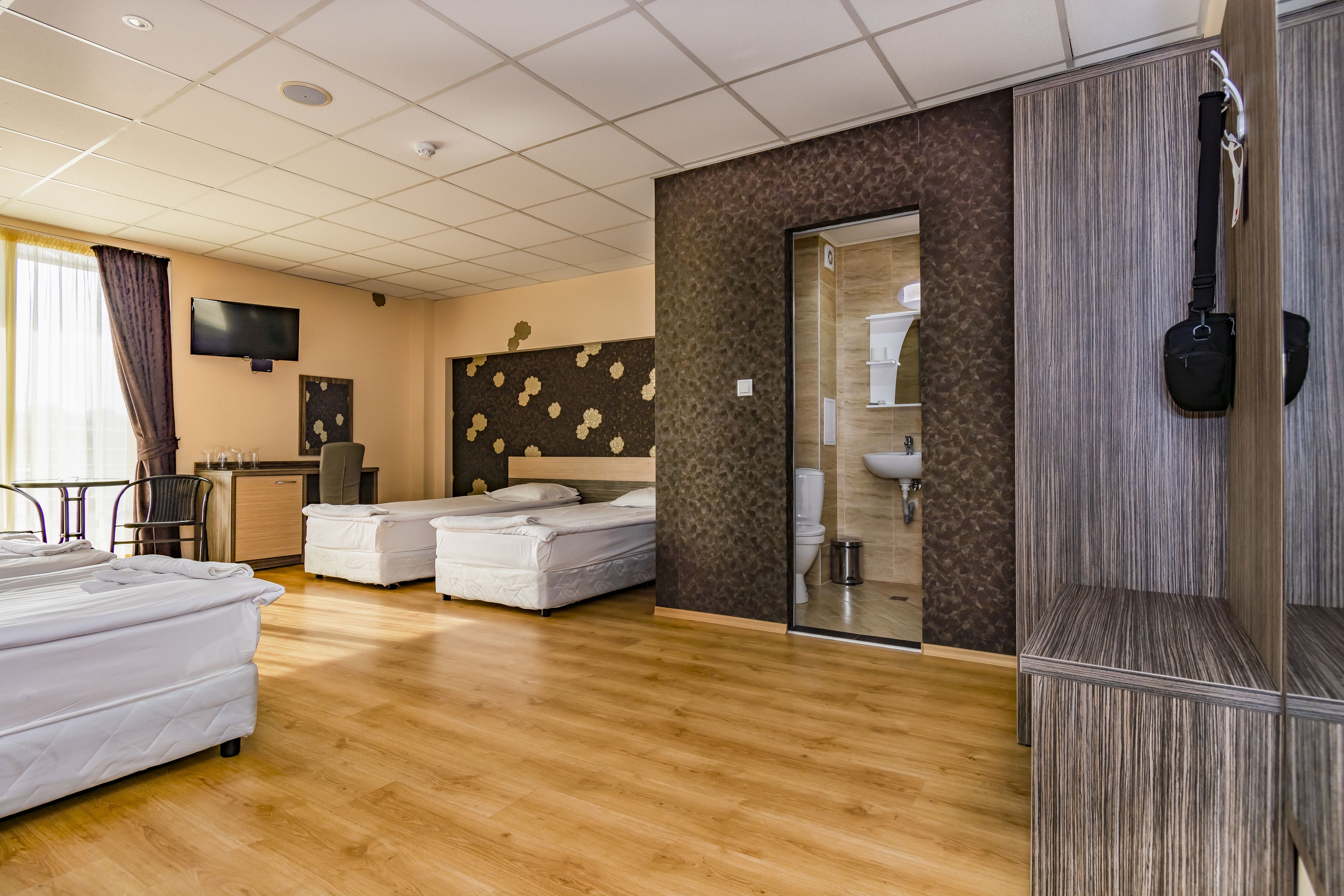 Quarto de hotel com piso de madeira, uma cama, uma escrivaninha, uma televisão e banheiro com pia e vaso sanitário.