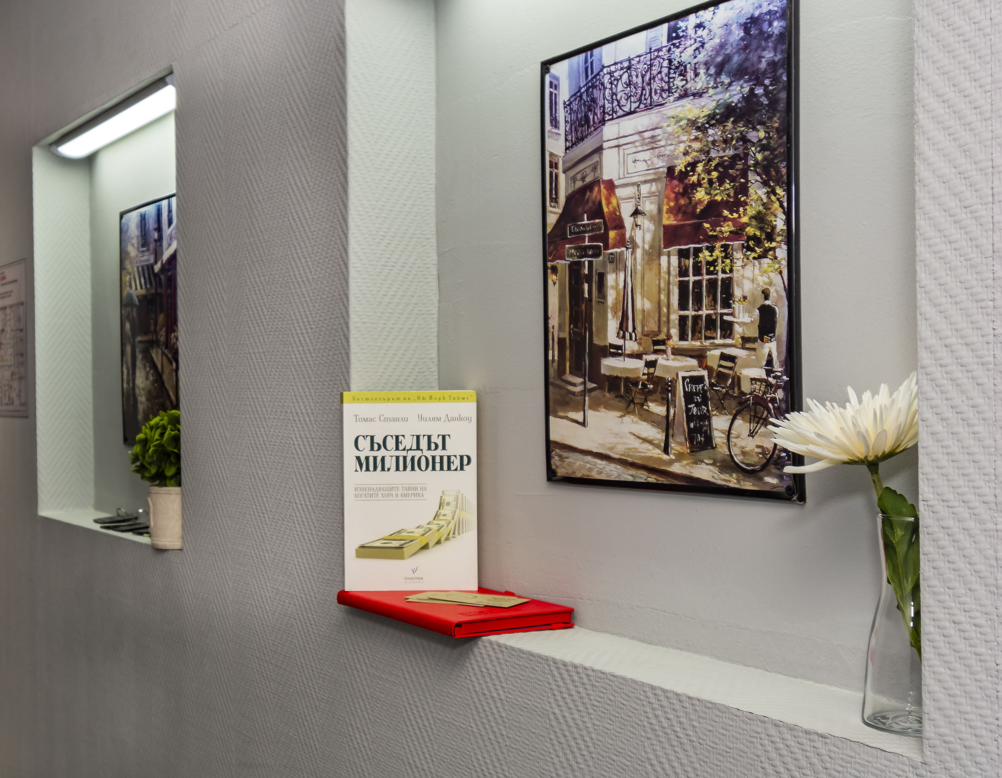 Een gang met een schilderij van een straatbeeld, een boek getiteld 'СБЕЖТ МИЛИОНЕР' op een rode boekenstandaard, en een vaas met een witte bloem.