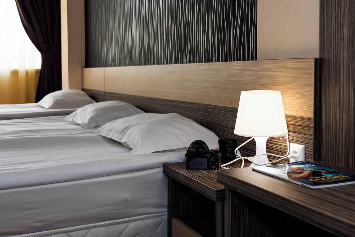 A hotel room with two beds, a wooden headboard, a nightstand with a lamp, a camera, and a magazine.