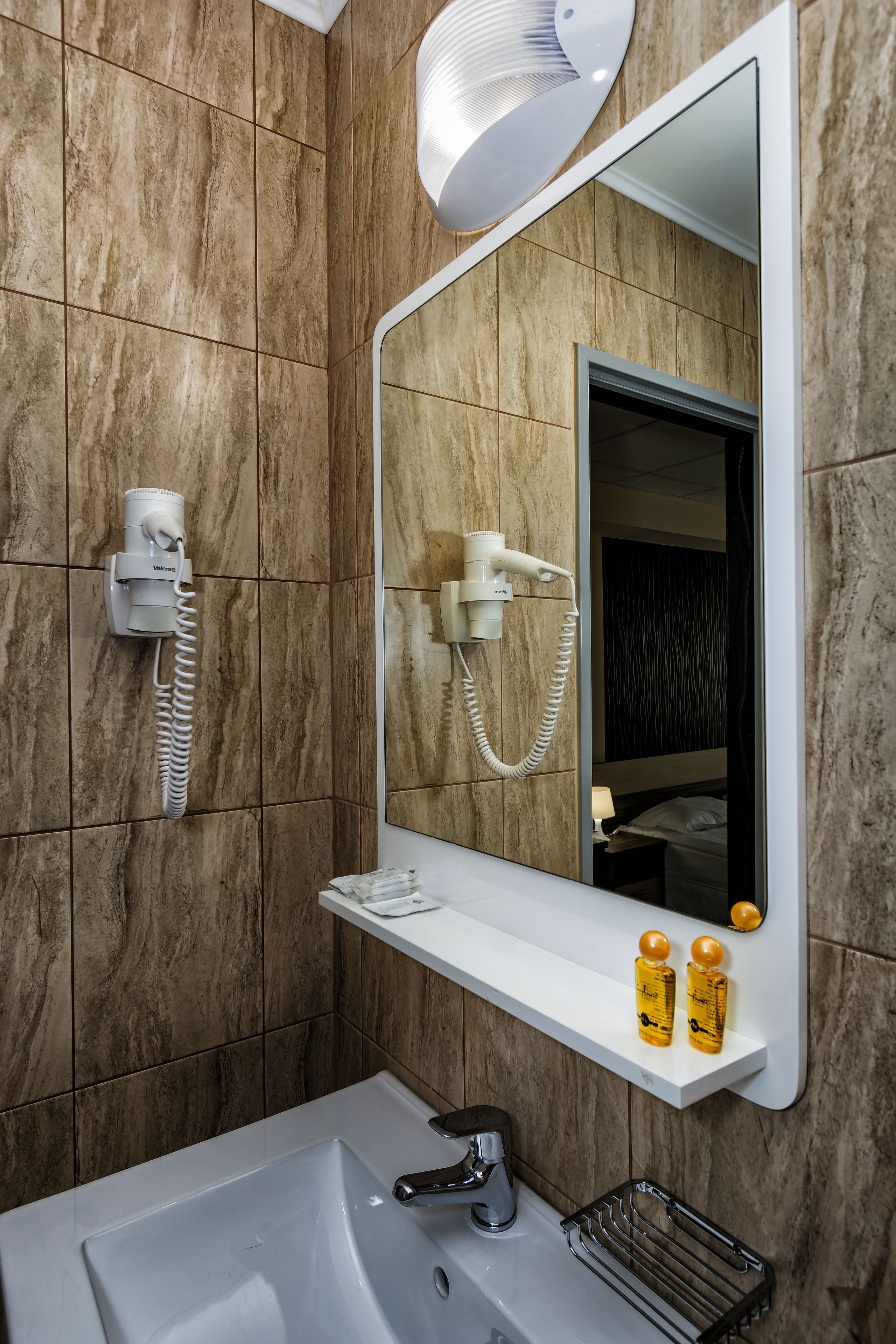 A bathroom with a marble wall, a white sink, a mirror, a hairdryer, and two bottles of lotion on a shelf.