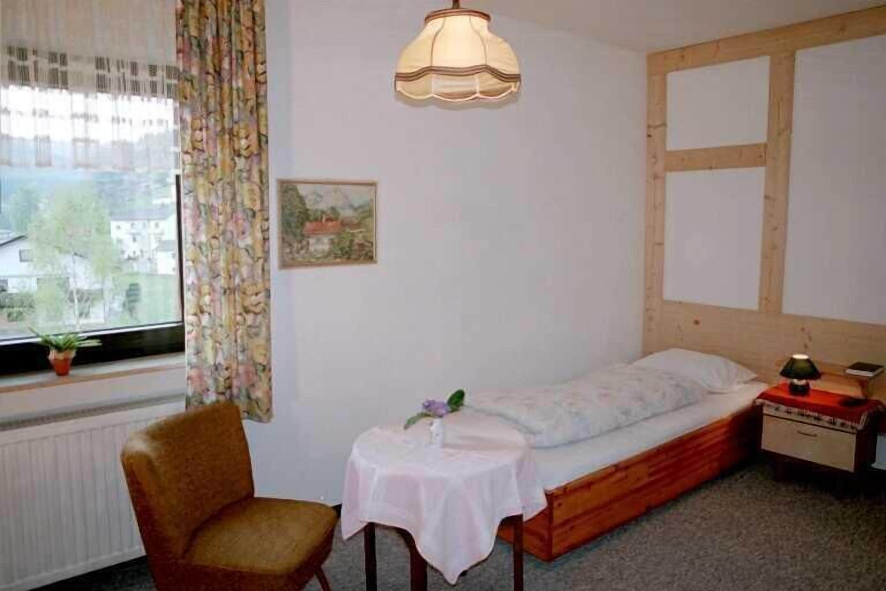 A small bedroom with a single bed, a chair, a small table, a lamp, and a window with curtains.