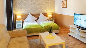 Une chambre d’hôtel avec un lit, deux tables de chevet avec des lampes, un téléviseur posé sur un meuble, un canapé et une petite table avec une plante.