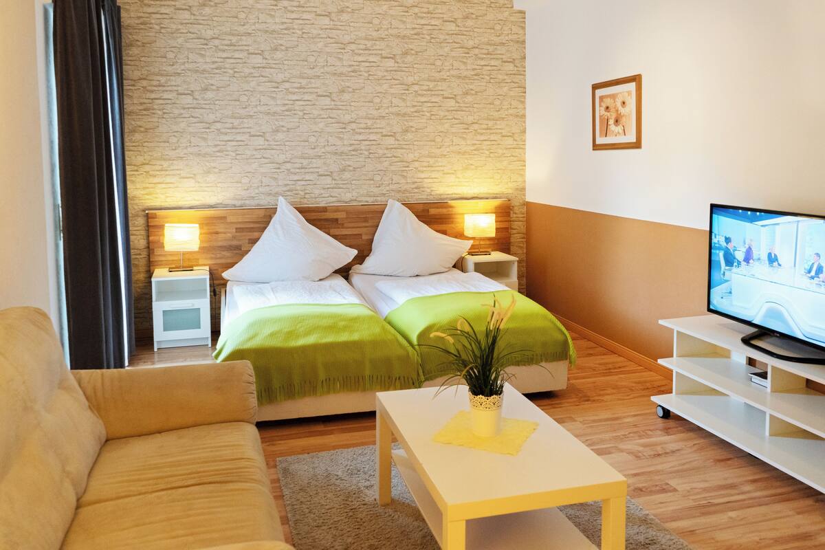 Une chambre d’hôtel avec un lit, deux tables de chevet avec des lampes, un téléviseur posé sur un meuble, un canapé et une petite table avec une plante.
