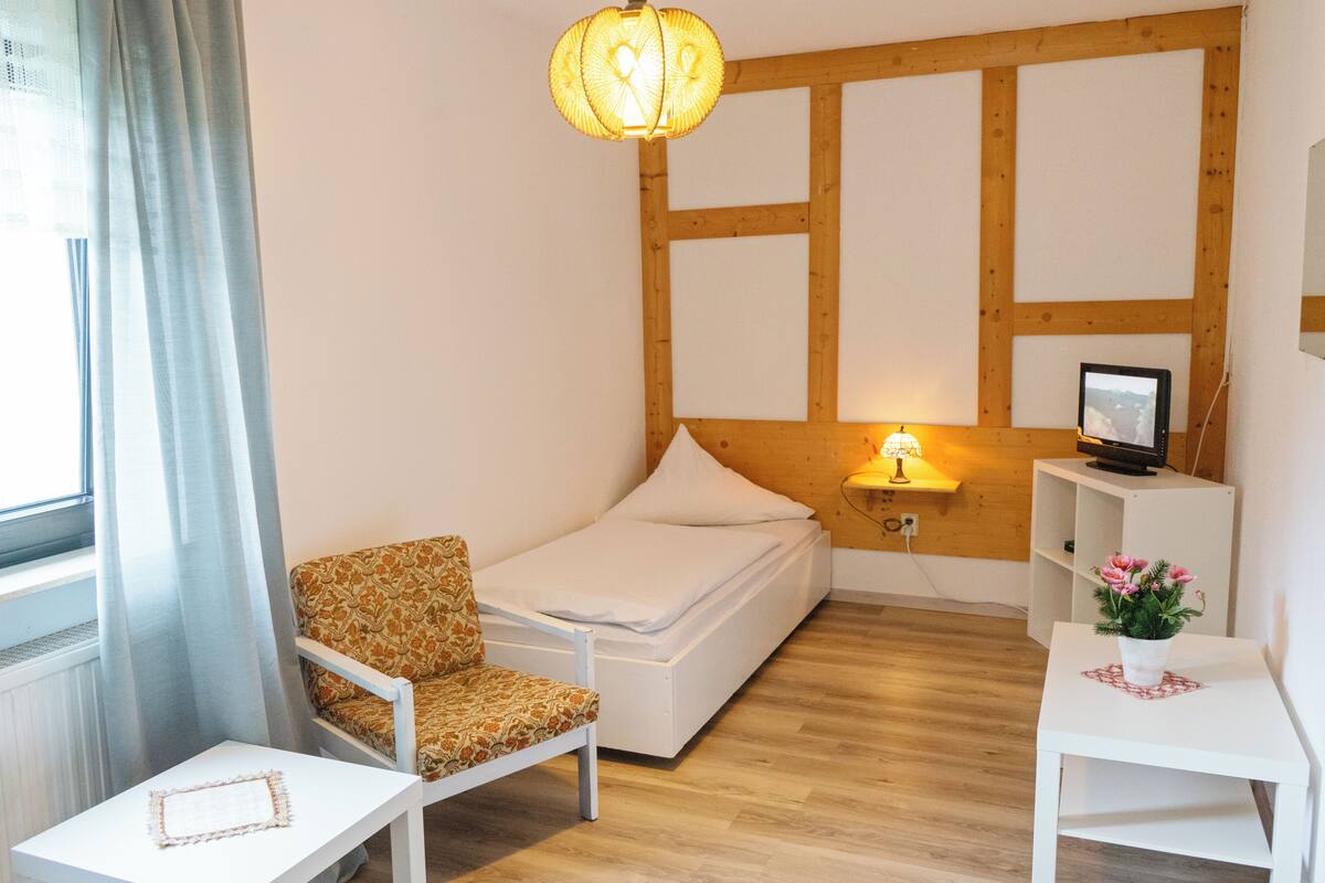 Une petite pièce bien éclairée, avec un lit, une chaise, une petite table, une lampe et un téléviseur posé sur une étagère.