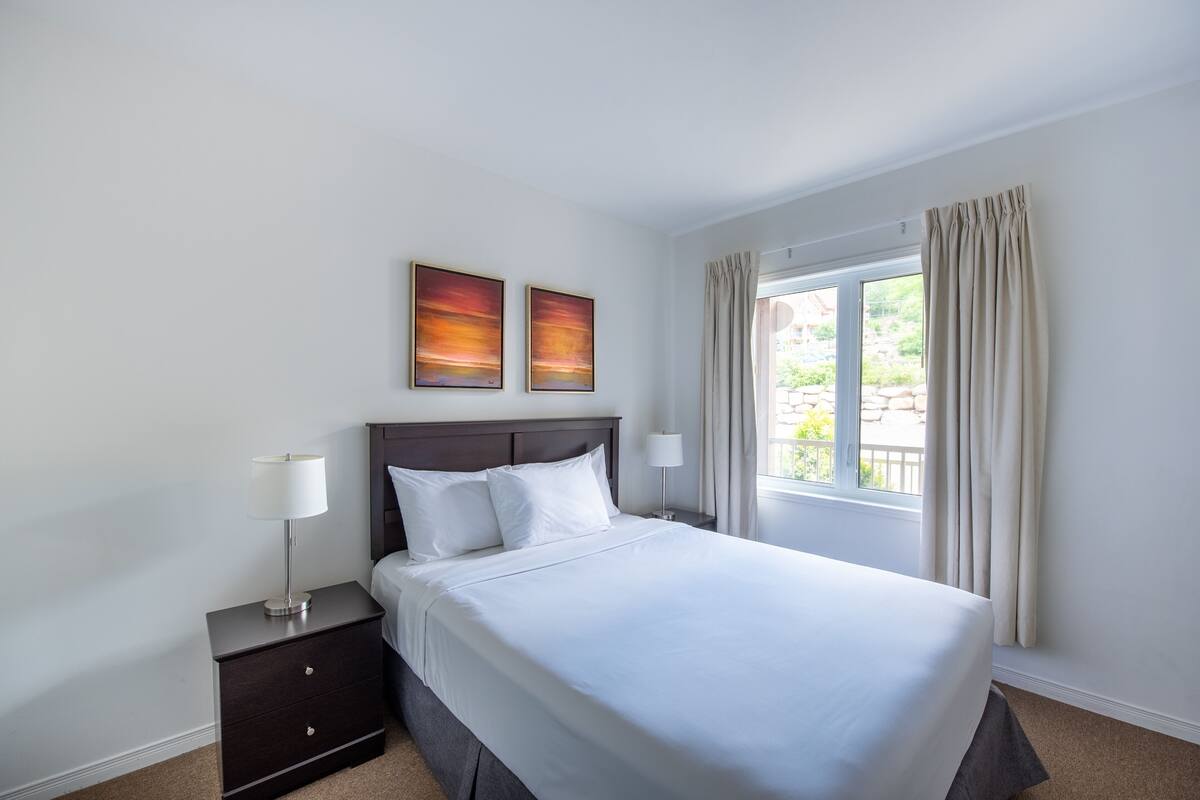 Una habitación de hotel con una cama, dos mesitas de noche, una ventana con cortinas y dos cuadros colgados en la pared.