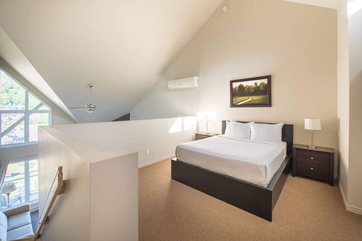 Un dormitorio con cama, mesitas de noche, ventilador de techo, aire acondicionado y ventana con vista a zonas verdes.