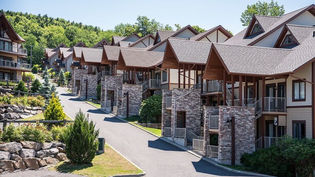 Une rangée d’immeubles modernes à plusieurs étages, dotés de façades en pierre et de balcons en bois, se dressant sur un fond de verdure luxuriante et d’un ciel dégagé.