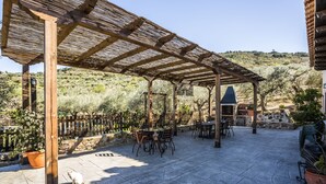 Een terras buiten met een pergola, stenen vloer en een eethoek met tafels en stoelen.