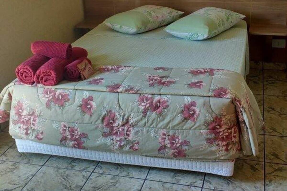 Een bed met bloemenbedekking, twee kussens en opgerolde handdoeken op een betegelde vloer.