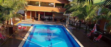 Una piscina rettangolare con un bordo in legno, una zona relax coperta con tavoli e sedie, e un edificio a due piani con tetto rosso sullo sfondo.