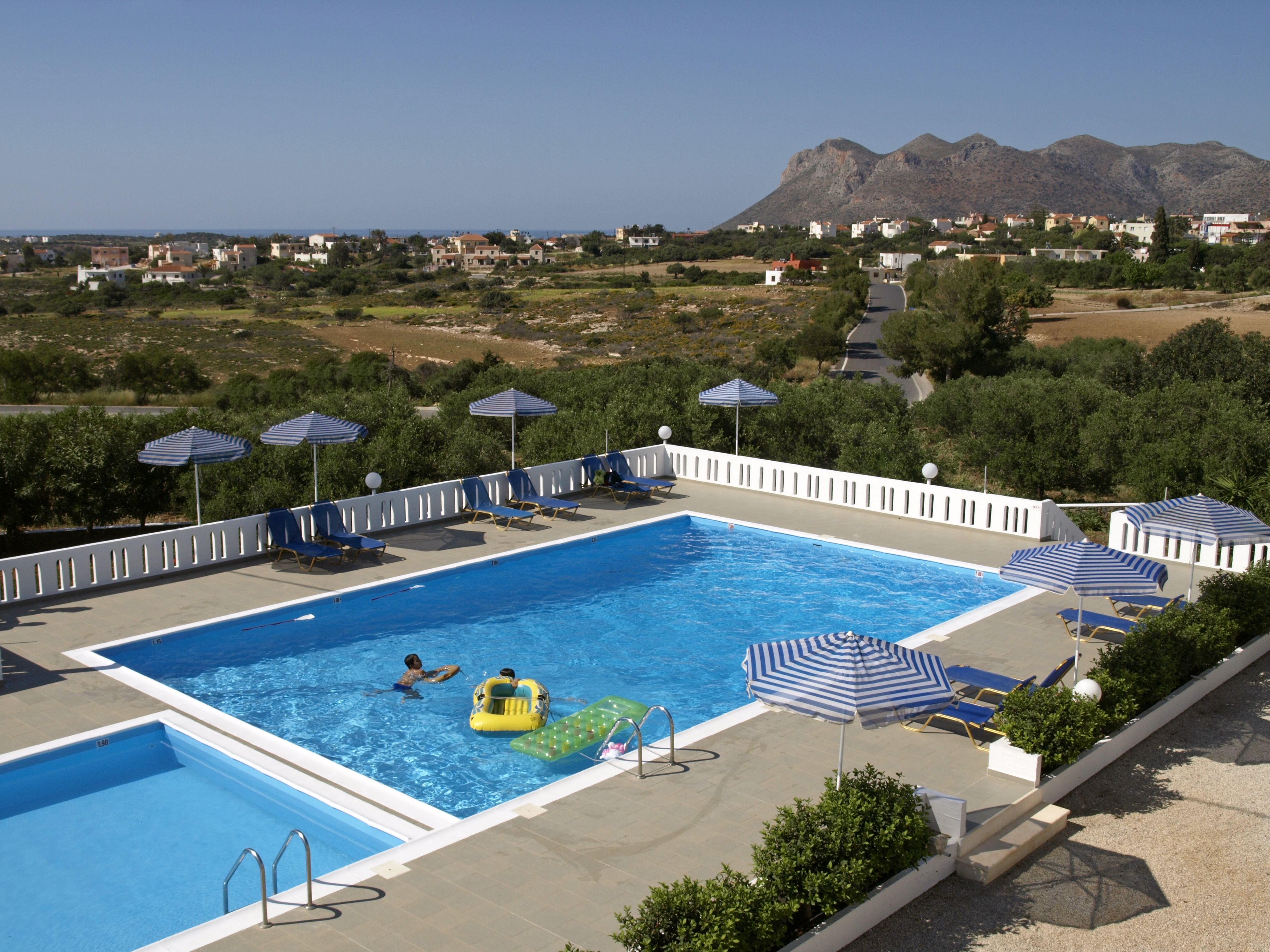 Piscina con sillas de descanso y sombrillas, rodeada de follaje y con vista a casas y montañas a lo lejos.