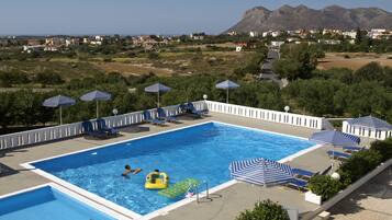 Piscina con sillas de descanso y sombrillas, rodeada de follaje y con vista a casas y montañas a lo lejos.