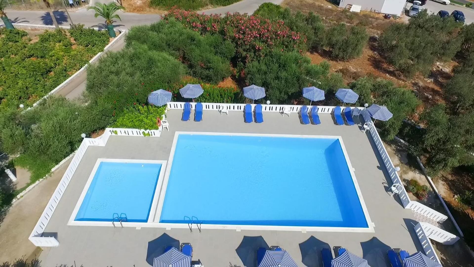 Una piscina en la azotea con dos piscinas contiguas, rodeada de sillones de descanso y sombrillas, con follaje y una carretera al fondo.