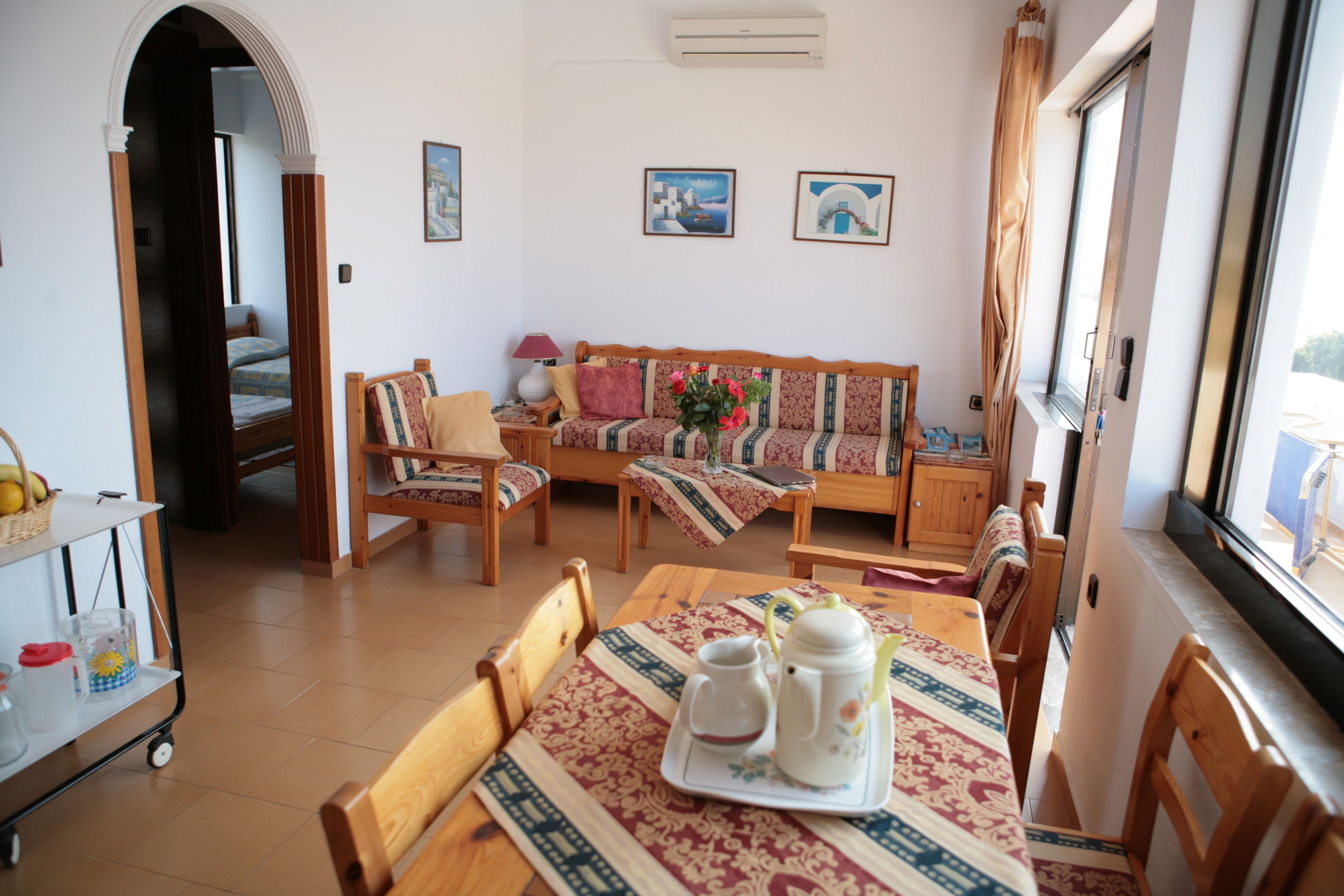 A cozy living room with a sofa, chairs, a dining table with a tray of tea set, and a window with curtains.