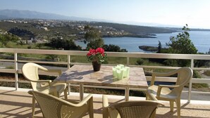 A table with chairs and a vase of flowers on a balcony overlooking a coastal landscape with houses and mountains in the distance. - Michalis Villas (Chania)