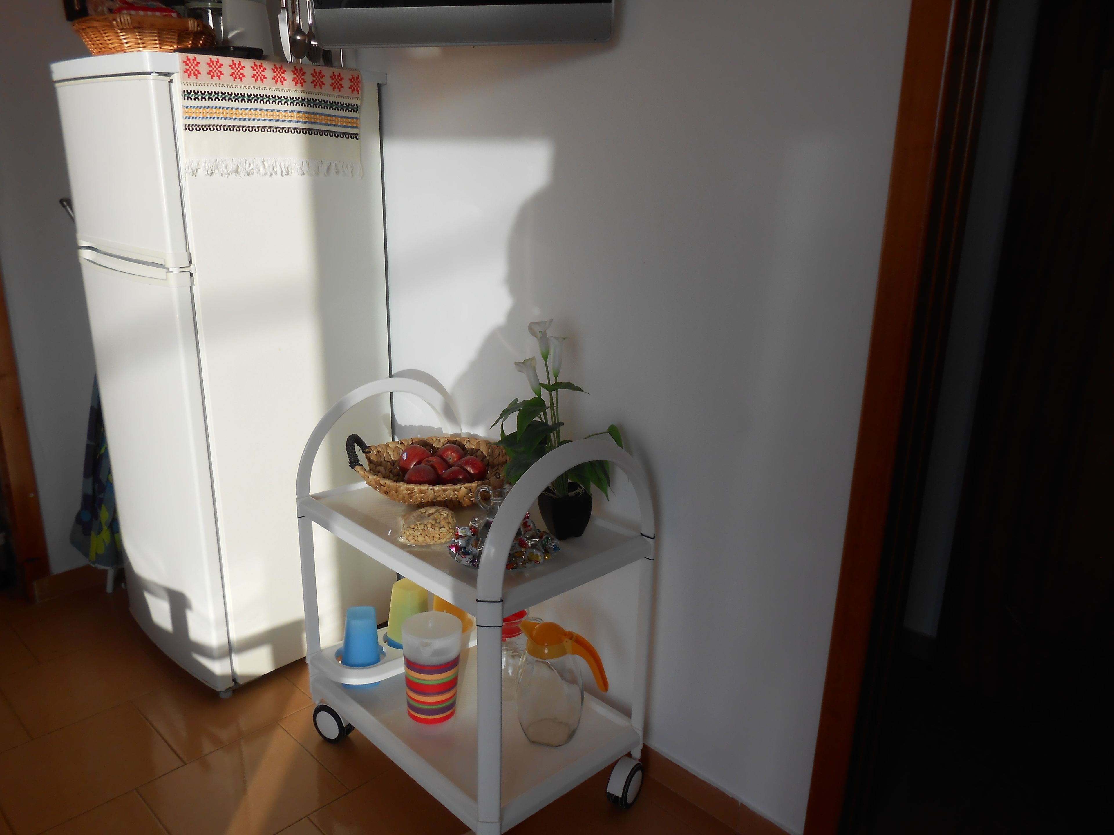 A white refrigerator with decorative patterns, a white cart with a basket of red apples, a potted plant, and a striped cup on a tiled floor.