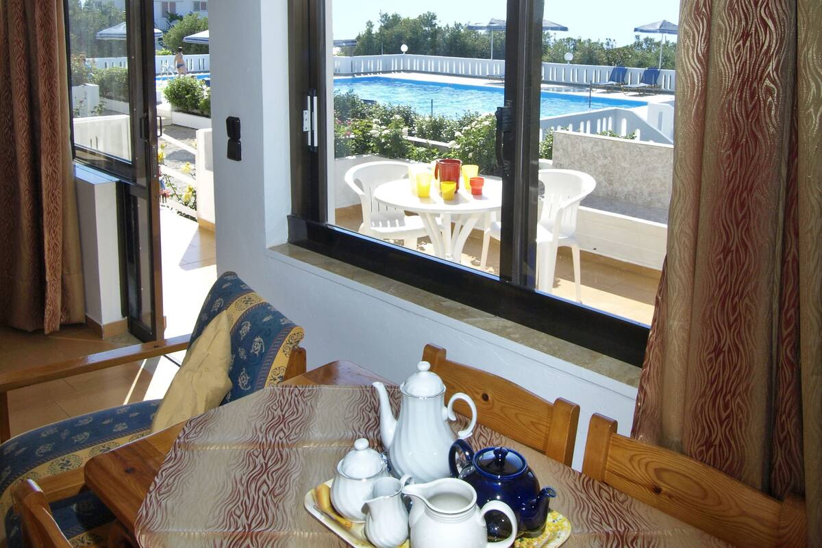 Comedor con una mesa preparada para tomar té, vista a una piscina y mobiliario exterior, además de un ventanal con cortinas.