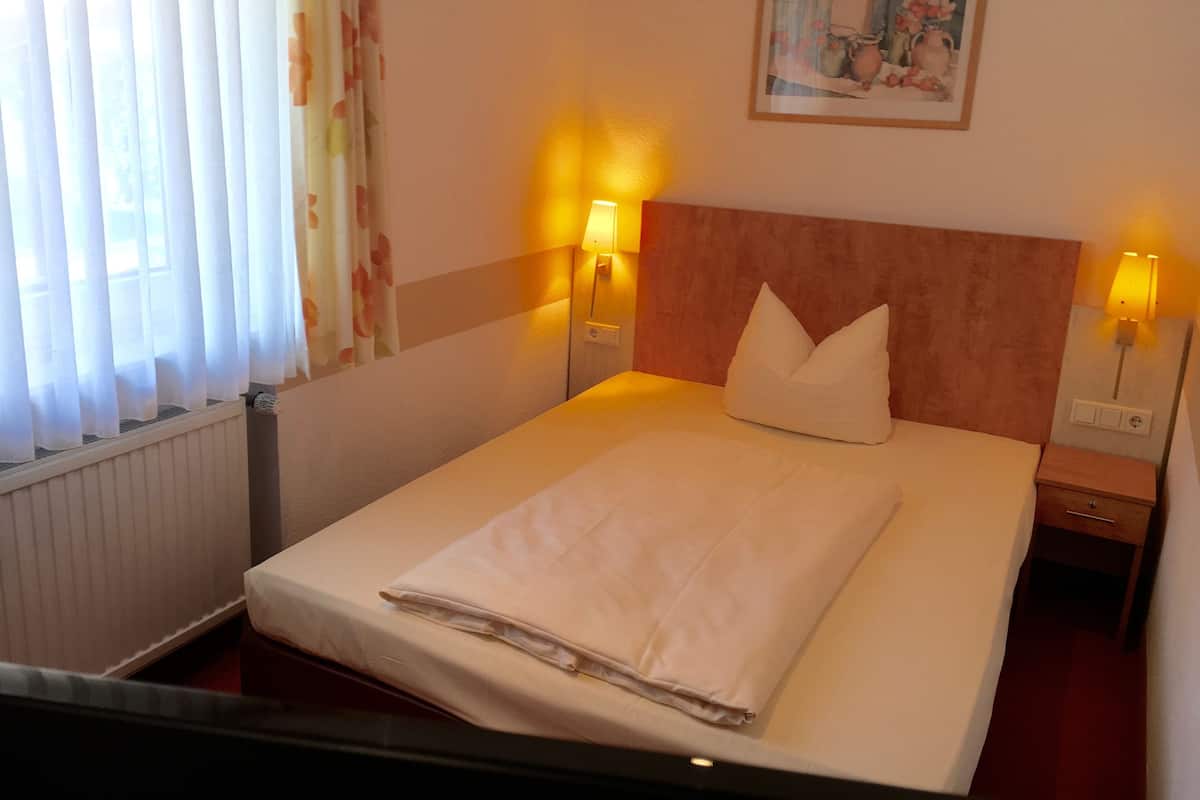 A hotel room with a neatly made bed, two bedside lamps, a headboard, a radiator, and a painting on the wall.