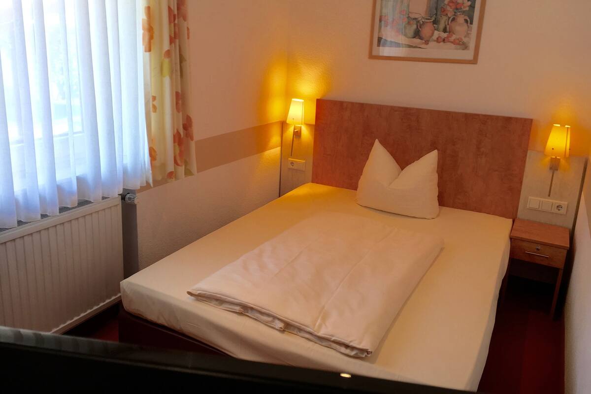 A neatly made bed with a white comforter and pillows, two bedside lamps, a framed picture on the wall, and a window with curtains.