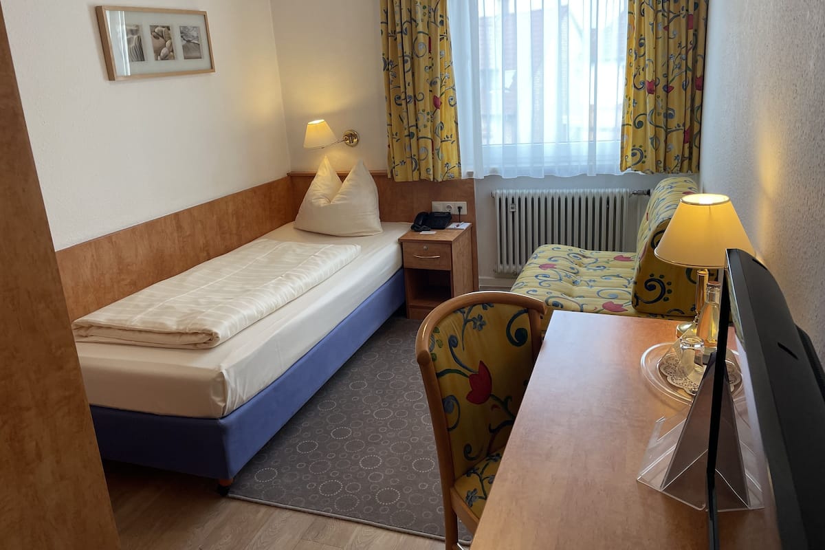 A hotel room with a single bed, a desk with a chair, a lamp, a picture on the wall, and a window with curtains.