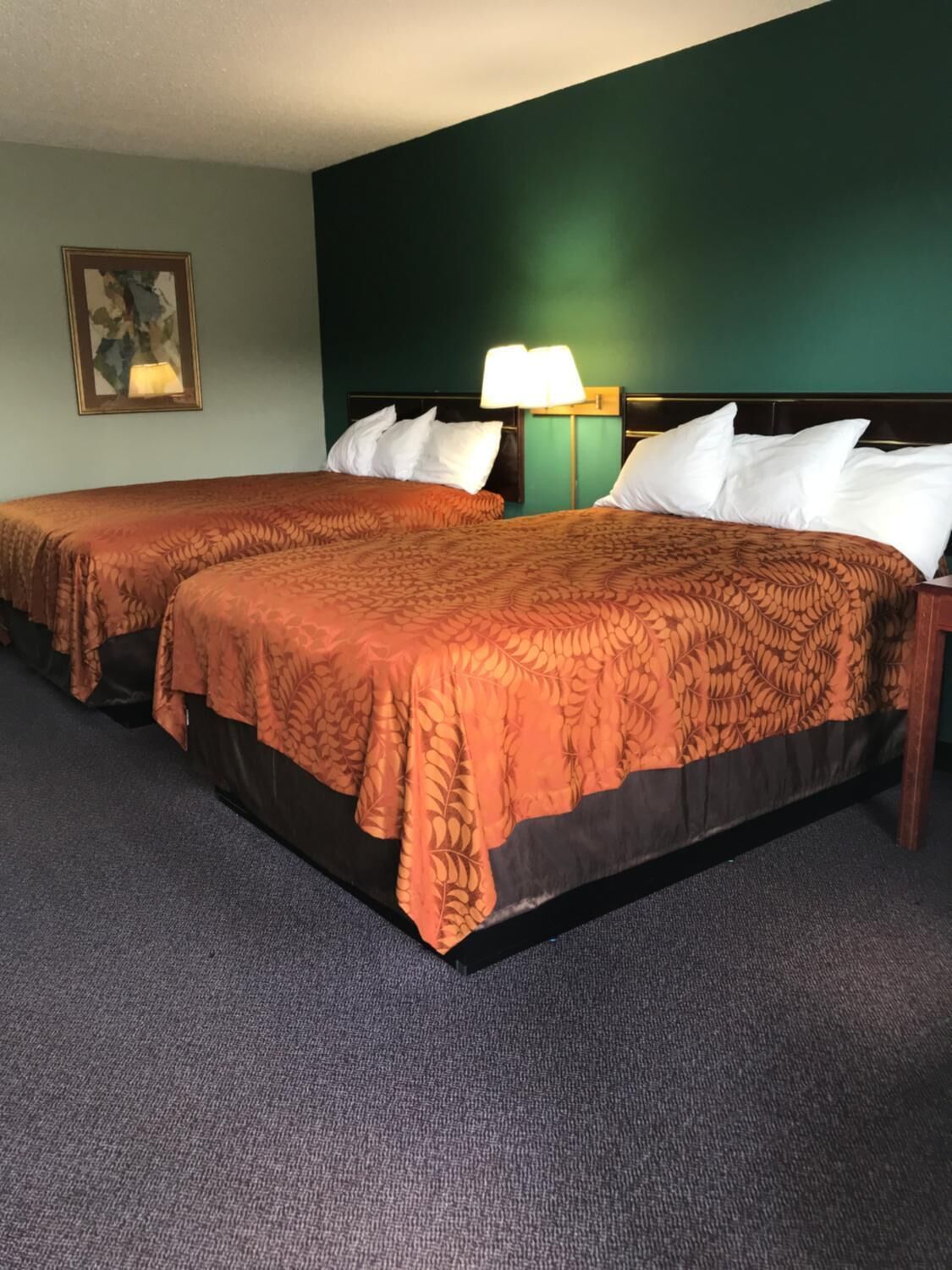 A hotel room with two beds, each with a patterned bedspread and white pillows. There is a nightstand with a lamp between the beds.