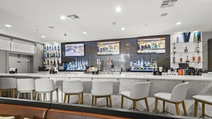 A bar area with white bar stools, a counter with a TV screen, and shelves of liquor bottles. - Holiday Inn Gonzales by IHG (Gonzales)