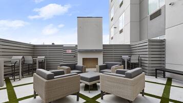 Uma área de estar moderna ao ar livre, com móveis de vime, lareira e vista para um edifício ao fundo.