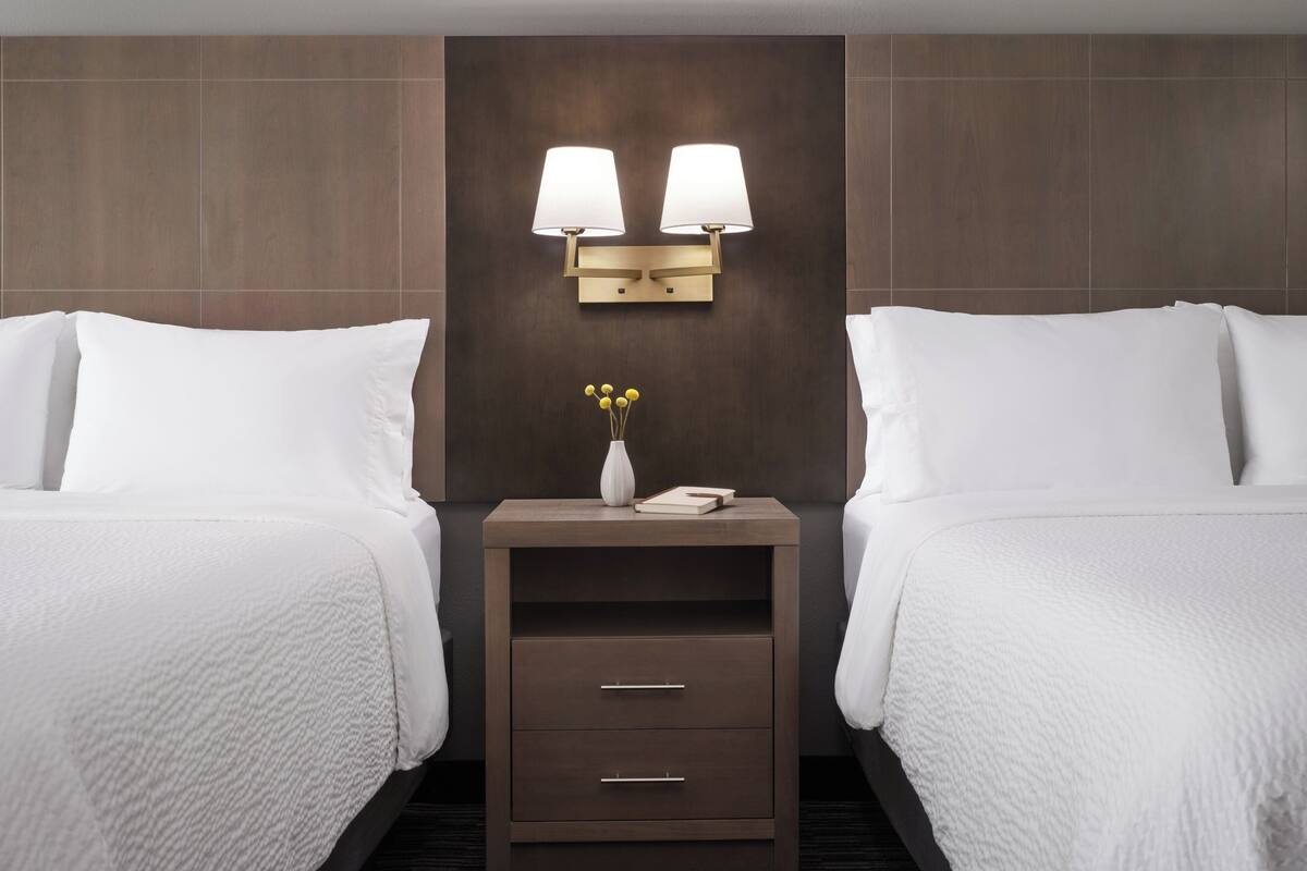 A hotel room with two beds, a nightstand with two wall-mounted lamps, a vase with yellow flowers, and a book.