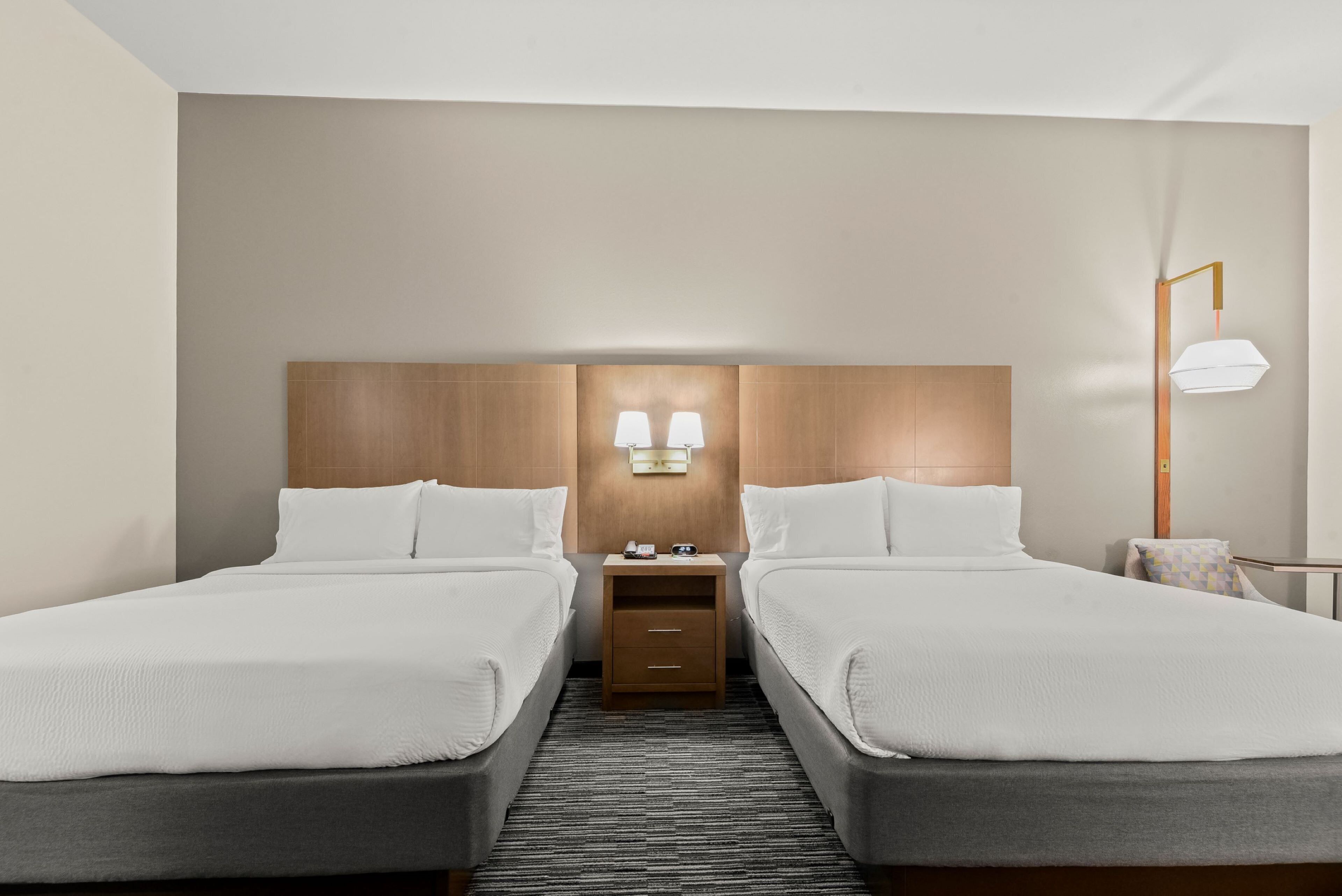 A hotel room with two beds, a wooden headboard, a nightstand with a lamp, and a small table with a chair.