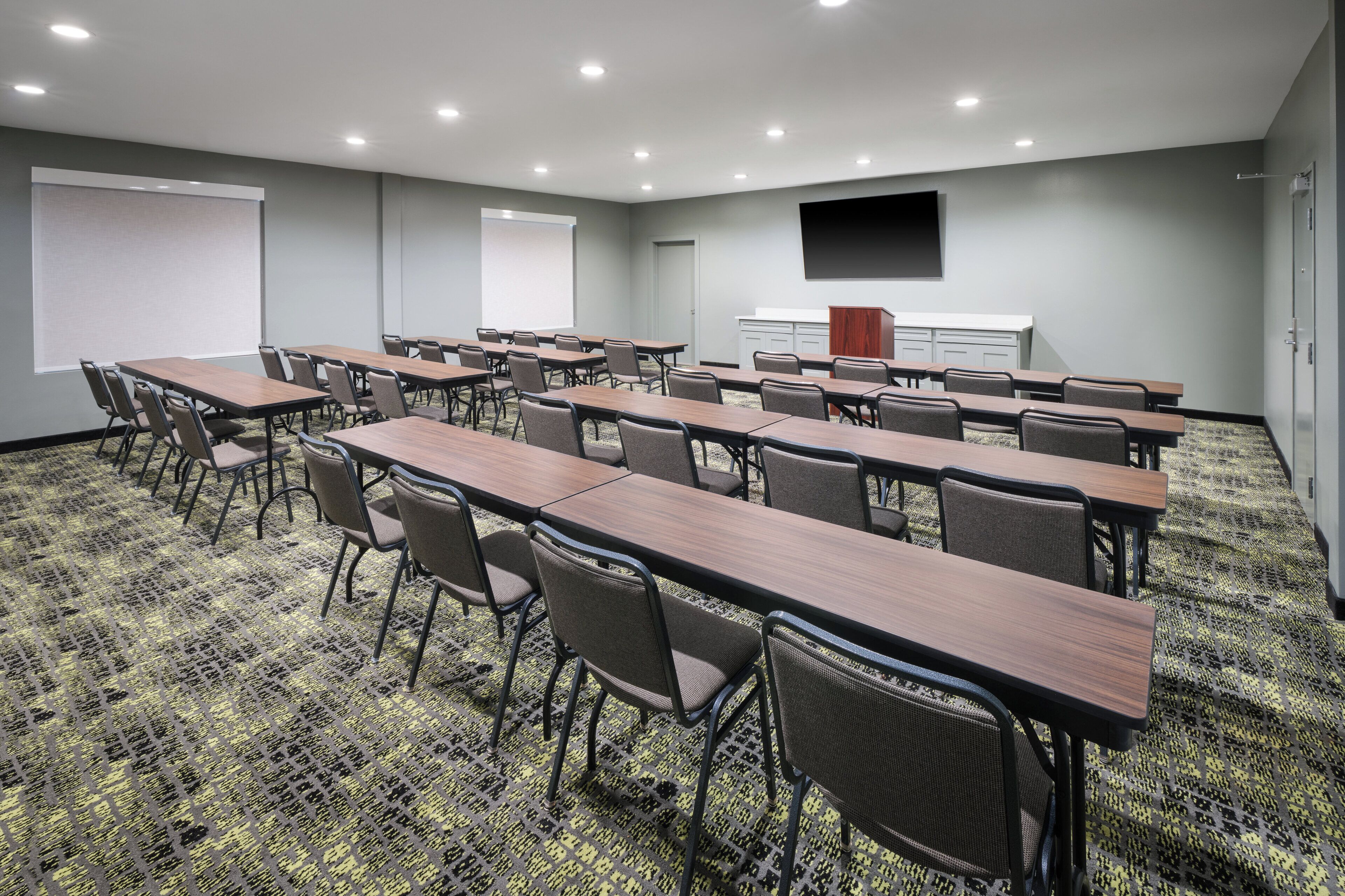 A conference room with multiple tables and chairs, a projector screen, a flat-screen TV, and a carpeted floor.