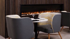 A modern dining area with a fireplace, round table, and grey upholstered chairs. - Holiday Inn Gonzales by IHG (Gonzales)