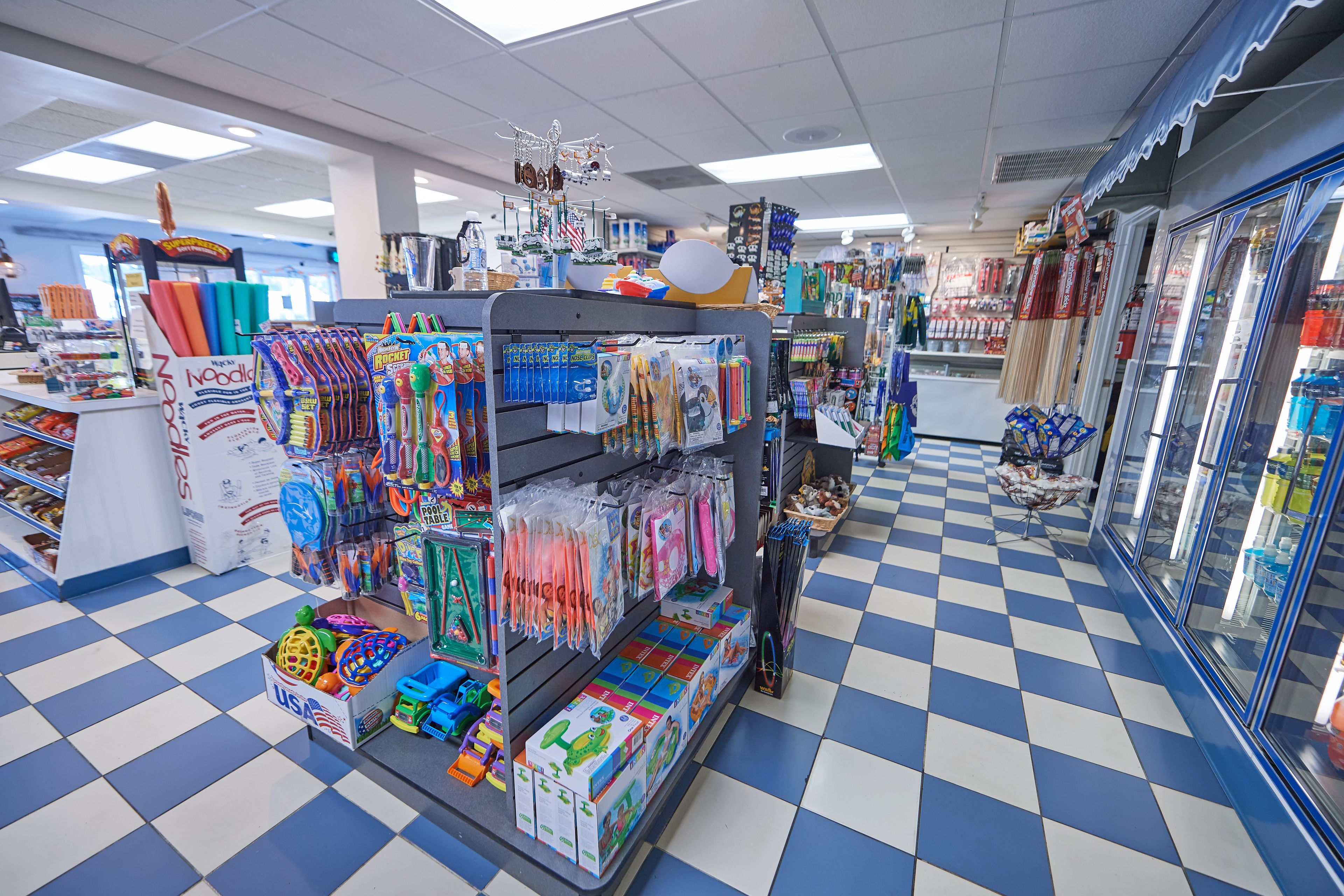 A store with a blue and white checkered floor, shelves stocked with toys and stationery, and a display case with various items.