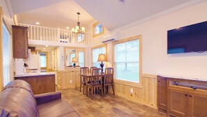 A spacious living area with a high ceiling, wooden flooring, a dining table with chairs, a sofa, and a television mounted on the wall. - Grey's Point Camp (Topping)
