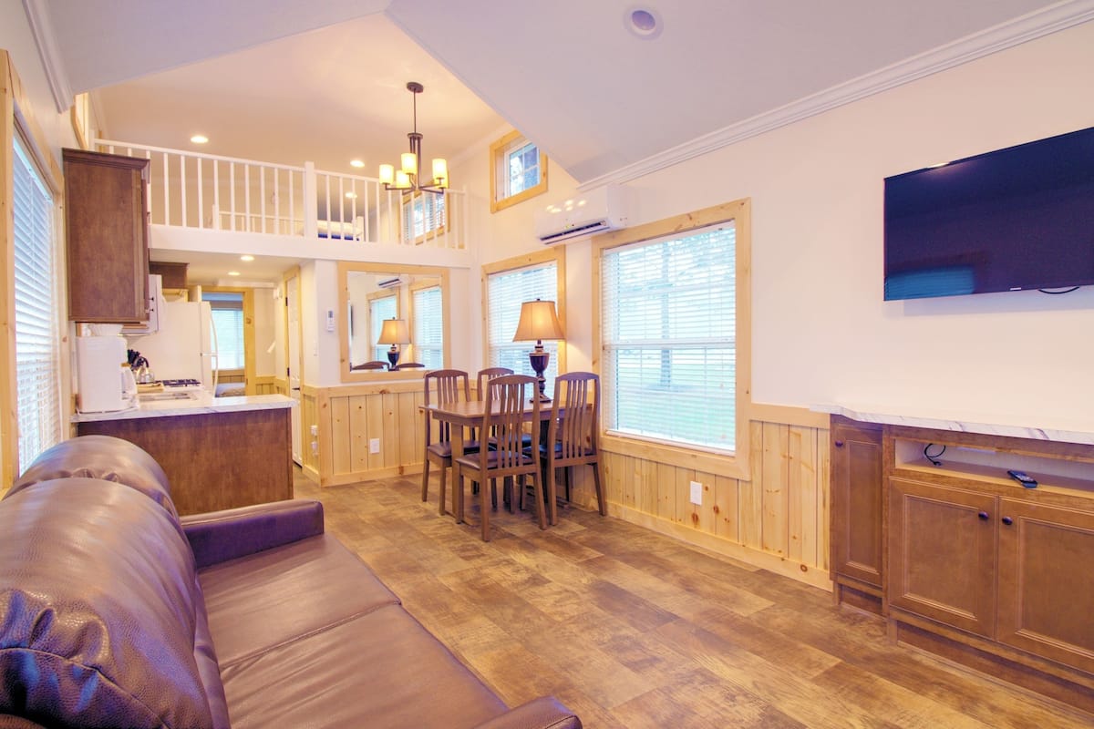 A spacious living area with a high ceiling, wooden flooring, a dining table with chairs, a sofa, and a television mounted on the wall.